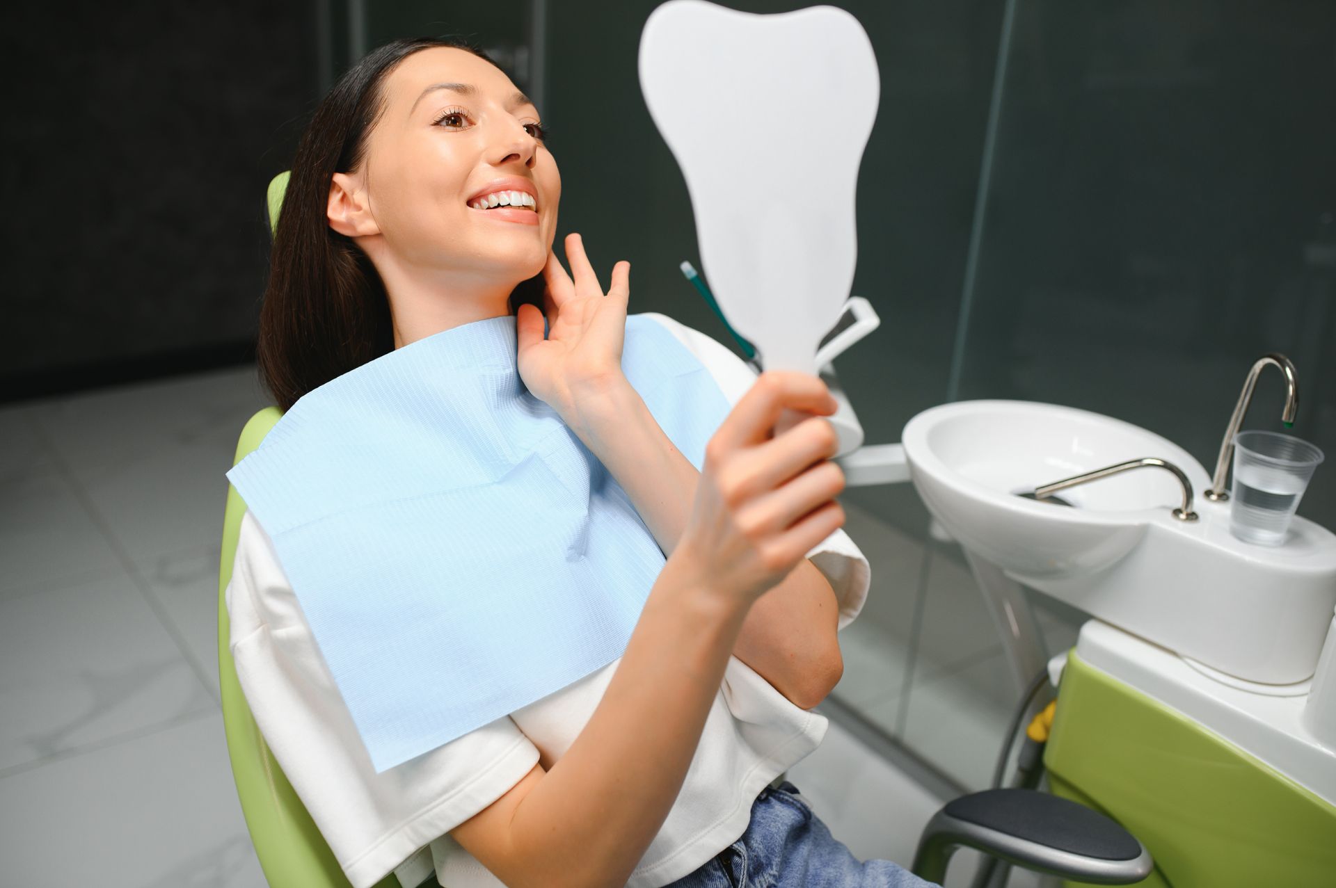 Female dental patient in Wilsonton Heights smiling and checking her teeth in a mirror after a cosmetic dentistry treatment, seated in a modern dental chair with a bib and visible dental sink nearby