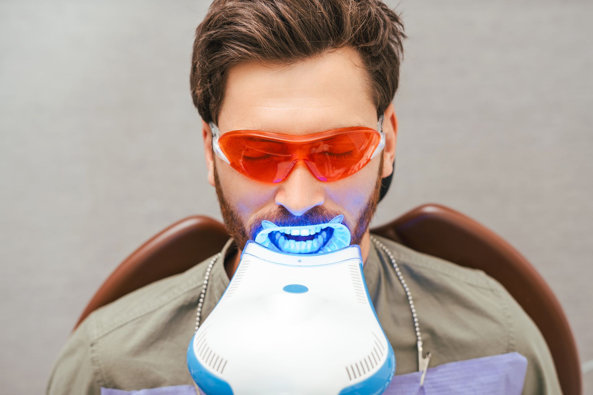 Man sitting in a dental chair wearing red goggles during a teeth whitening procedure with a special light scanner