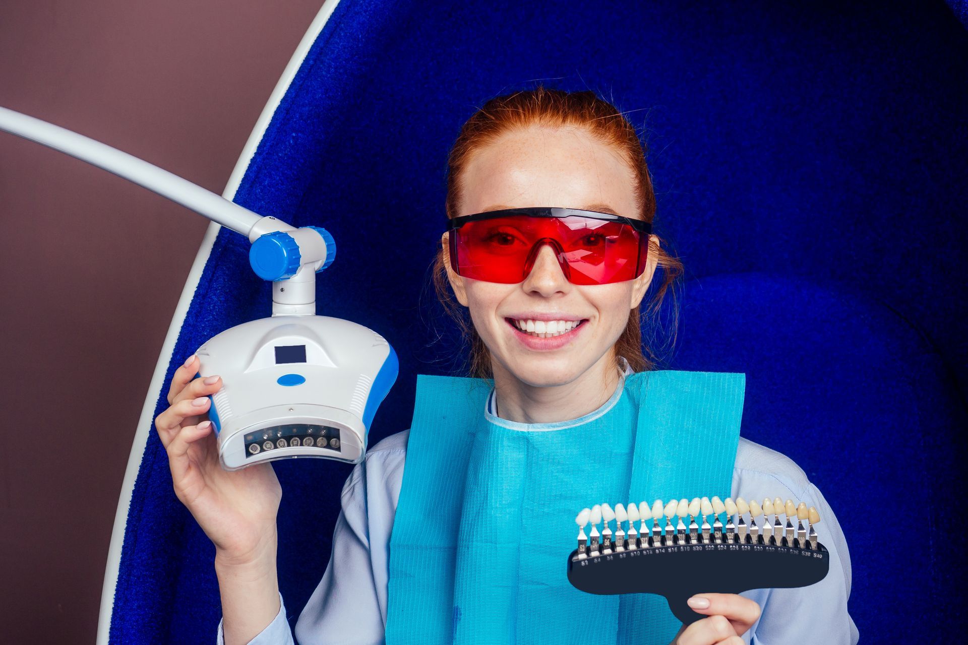 Cheerful young red-haired woman holding teeth shade guide and LED whitening machine, wearing protective glasses, ready for professional teeth whitening treatment in Wilsonton, QLD