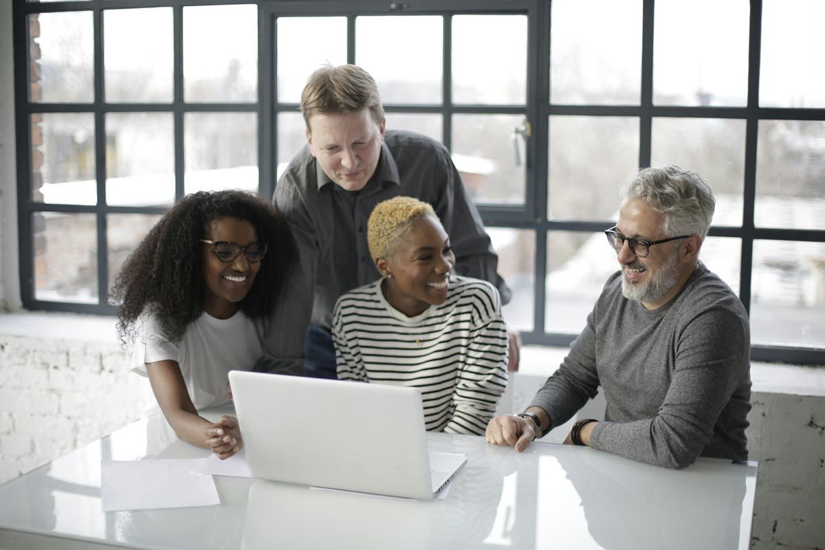 Four coworkers gathered around a laptop at a bright table in a modern office.
