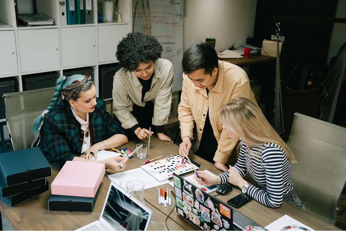 Four people collaborate around a table with papers, laptops, and craft materials in an office.