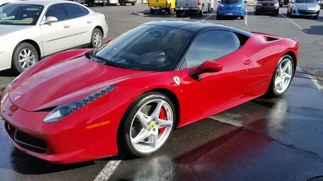 Red Ferrari sports car parked on a wet lot, with other cars in the background.