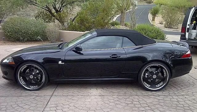 Black convertible sports car parked on a driveway, side view, with a black soft top and custom rims.