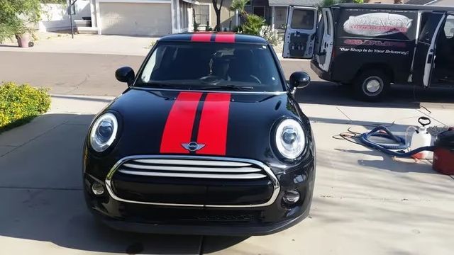 Black Mini Cooper with red racing stripes parked in a driveway, front view.