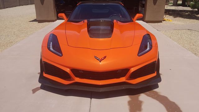 Orange Chevrolet Corvette parked on a driveway, front view with black hood accents.