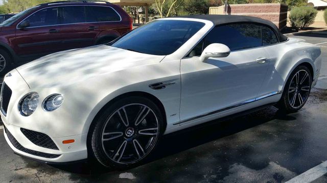 White Bentley convertible parked outdoors on a sunny street, with a maroon SUV behind it.