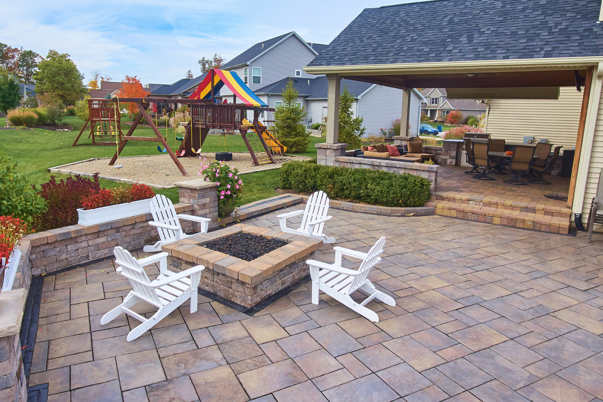 A patio with a fire pit and chairs and a playground in the background.