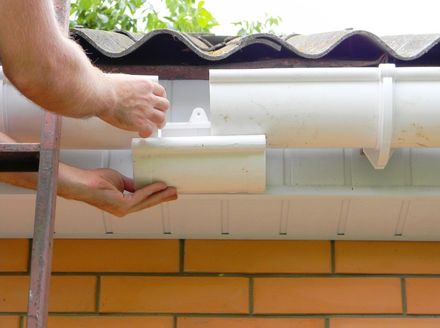 Person installing white gutter downspout on a brick building.
