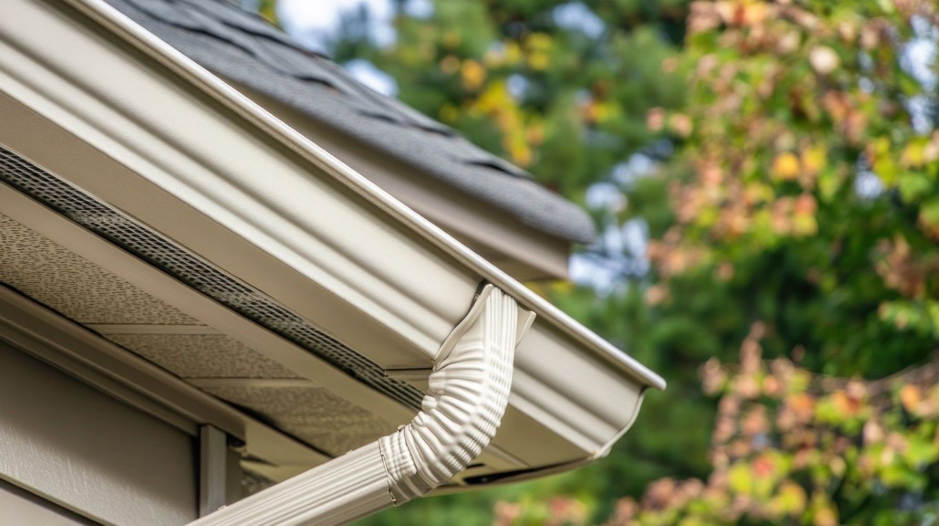 Beige rain gutter and downspout on a house, angled against a backdrop of green and yellow trees.