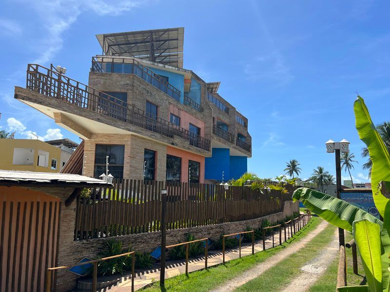 Prédio de vários andares com fachada colorida, varanda e painéis solares. Cerca marrom e caminho gramado na frente.