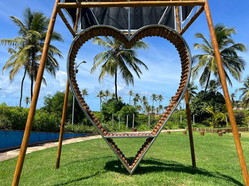 Grande escultura em forma de coração em um parque, emoldurada por palmeiras e um céu azul.