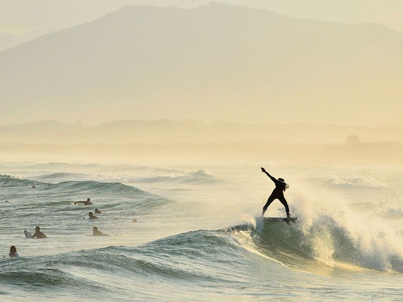 Surfista se equilibrando em uma onda, braços estendidos, perto de outros surfistas. Montanhas ao fundo, envoltas em névoa.