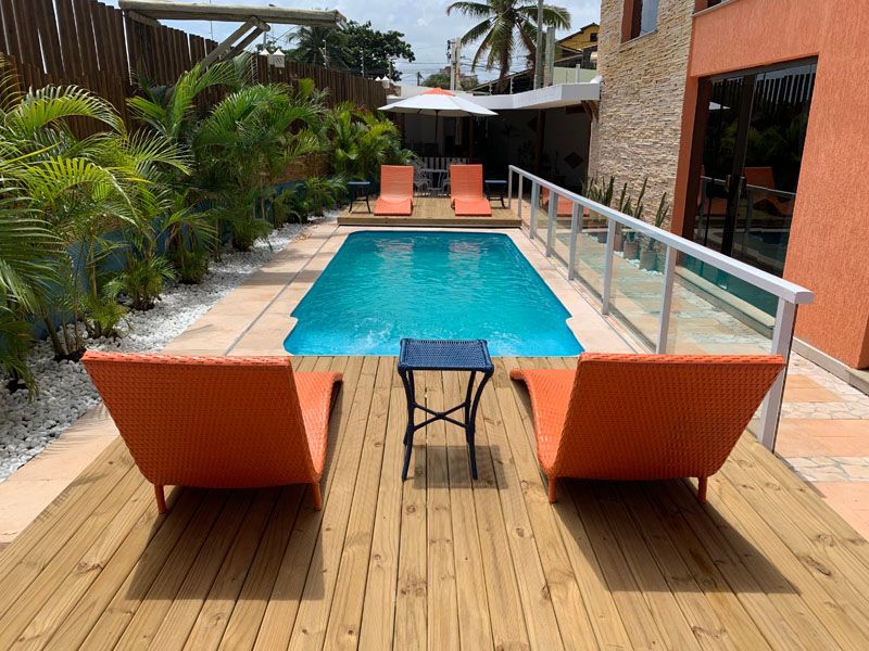 Área da piscina com espreguiçadeiras laranja, mesa pequena, piscina azul, deck de madeira e paisagismo.