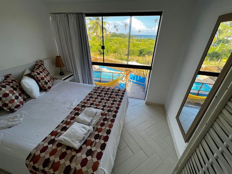 Quarto com cama de casal, manta estampada e uma grande janela com vista para a praia.