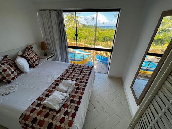Quarto com cama de casal, manta estampada e uma grande janela com vista para a praia.