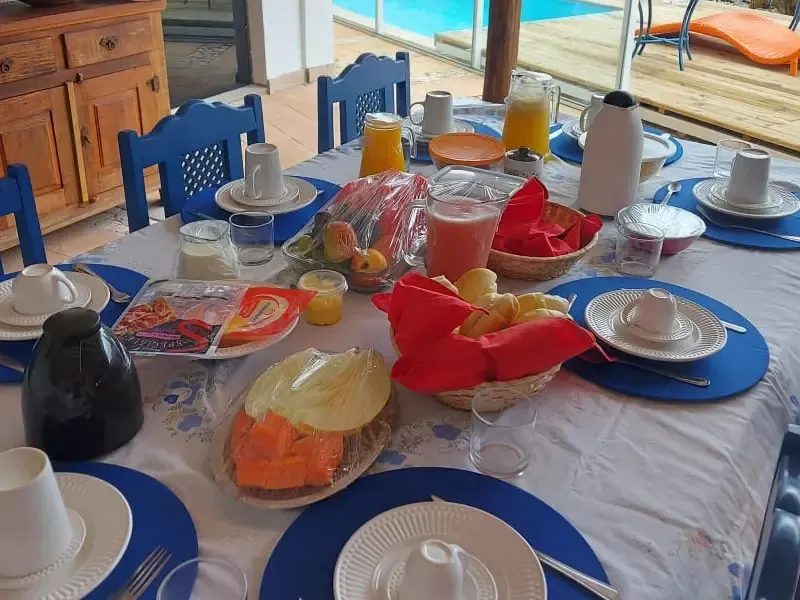 Mesa de café da manhã posta com alimentos, bebidas e louças. Inclui pão, queijo, suco, frutas e café. Jogos americanos azuis.