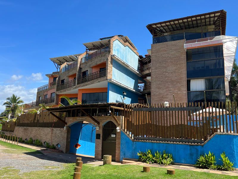 Prédio colorido de vários andares com seções azuis, laranjas e de tijolos, atrás de uma cerca de madeira, sob um céu azul.