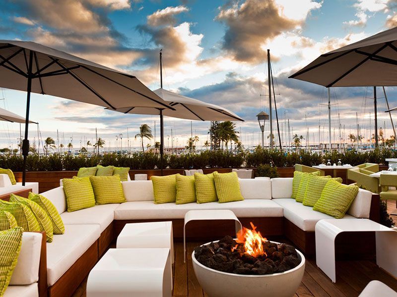 Lounge na cobertura com sofás brancos, almofadas verde-limão, lareira, guarda-sóis e vista para o porto ao pôr do sol.