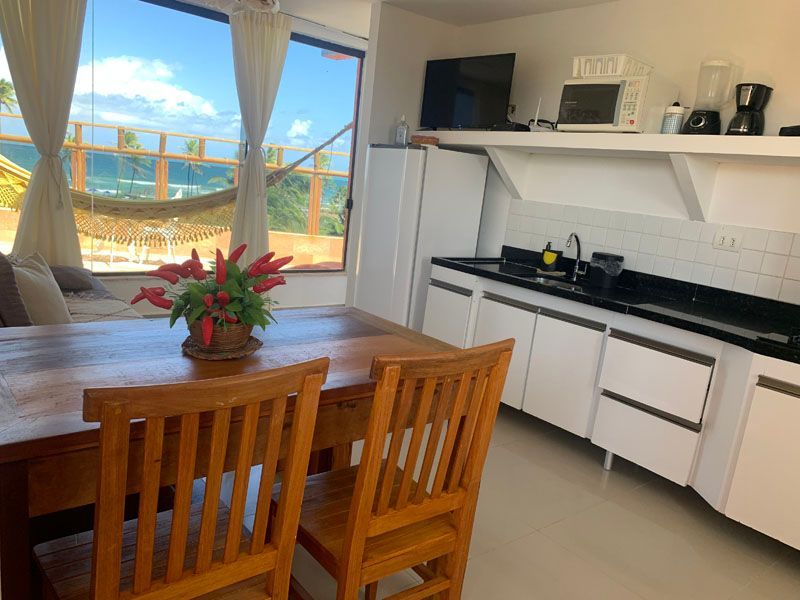 Cozinha e sala de jantar com vista para o mar. Mesa e cadeiras de madeira, armários brancos e uma rede.