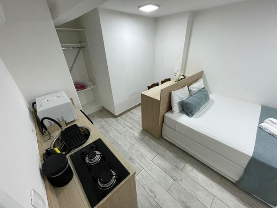 Quarto pequeno, com paredes brancas, cama, minicozinha e armário. Detalhes em madeira clara e piso cinza claro.
