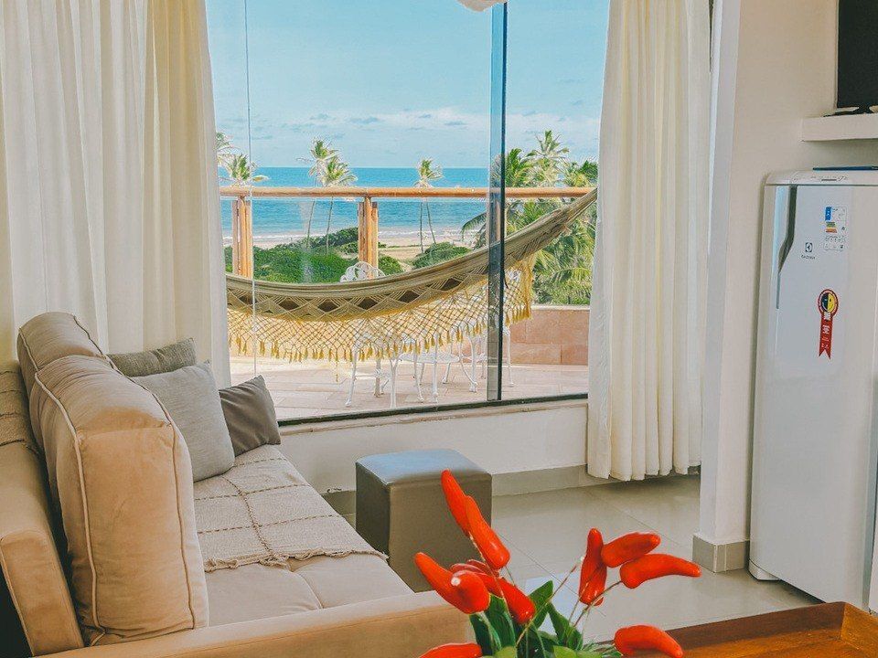 Sala de estar com vista para o mar: sofá bege, rede, cortinas brancas, pimentões vermelhos em um vaso e geladeira.