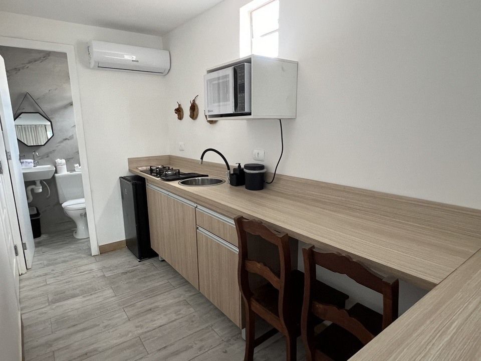 Pequena cozinha americana com armários de madeira clara, micro-ondas, pia e duas cadeiras. Banheiro visível ao fundo.