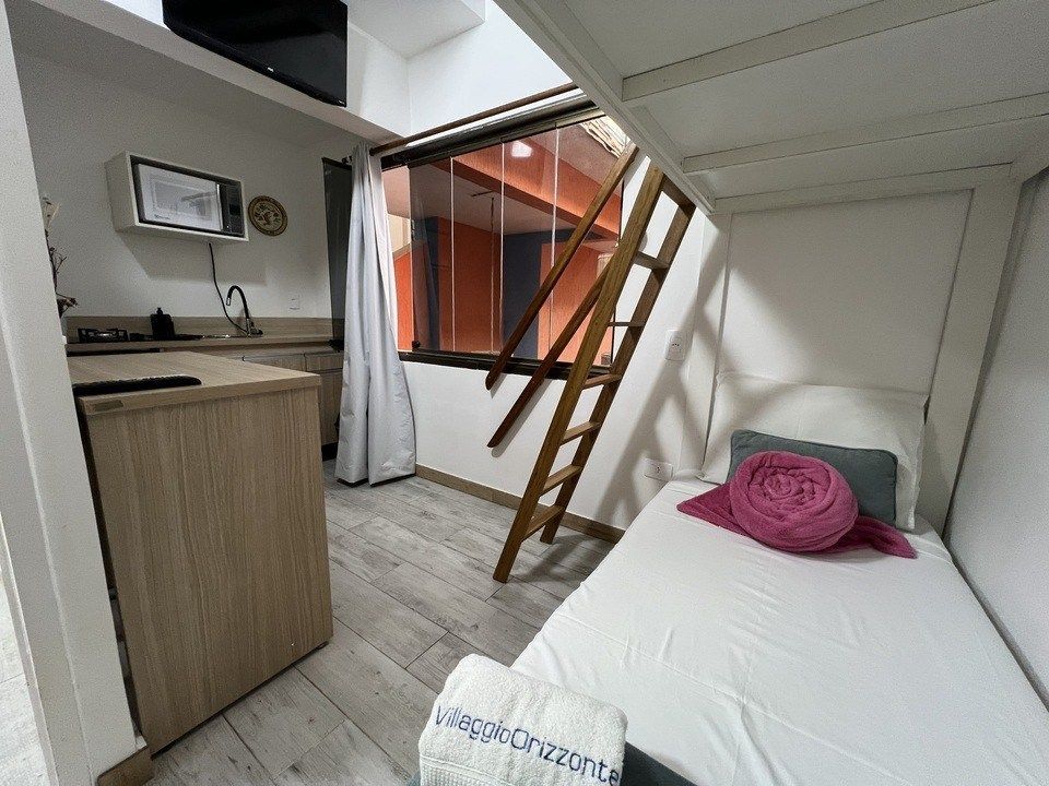 Quarto pequeno com beliche, balcão de cozinha, escada de madeira e janela. Tons de branco e madeira.