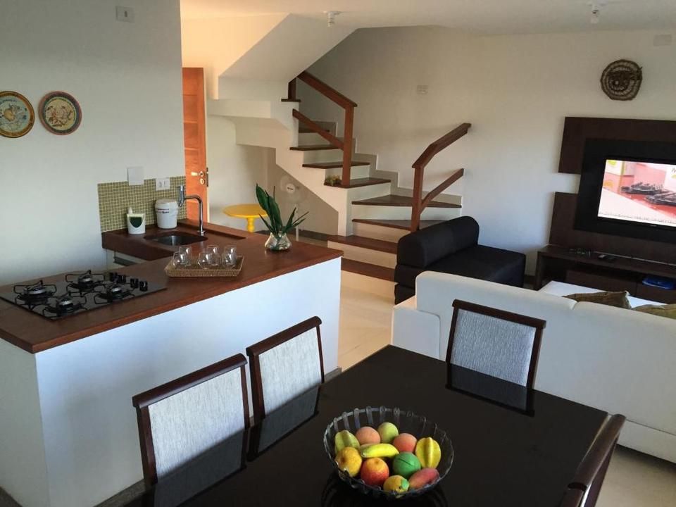 Espaço aberto com ilha de cozinha, mesa de jantar, escadas, sofá branco e TV em uma parede branca.