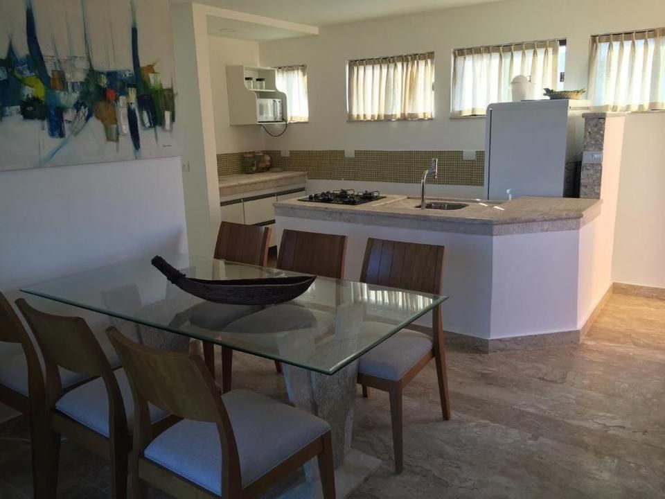 Sala de jantar com mesa e cadeiras de vidro, adjacente a uma cozinha com bancadas, eletrodomésticos e janelas.