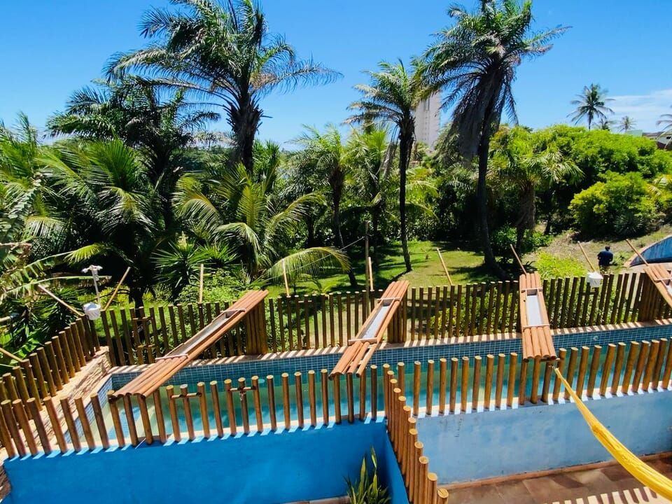 Área externa com piscinas azuis, cerca de bambu, palmeiras e vegetação exuberante sob um céu azul.