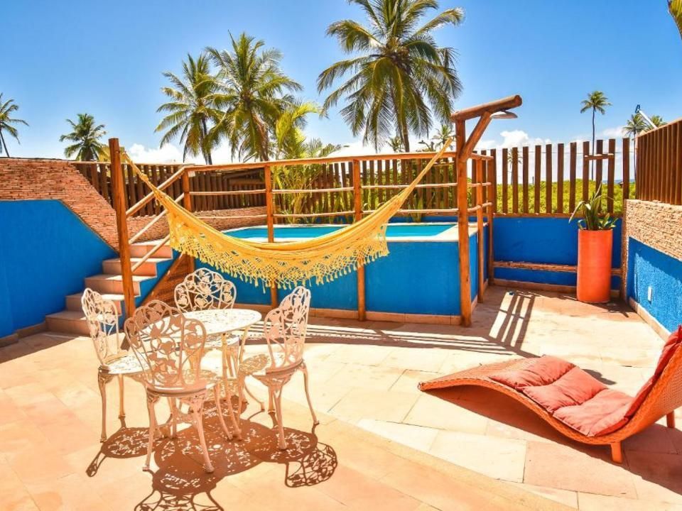 Pátio externo com piscina, rede, assentos e palmeiras; dia ensolarado.