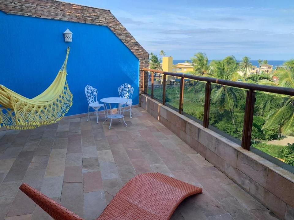 Terraço na cobertura com parede azul, rede amarela, mesa/cadeiras brancas e vista para o mar.