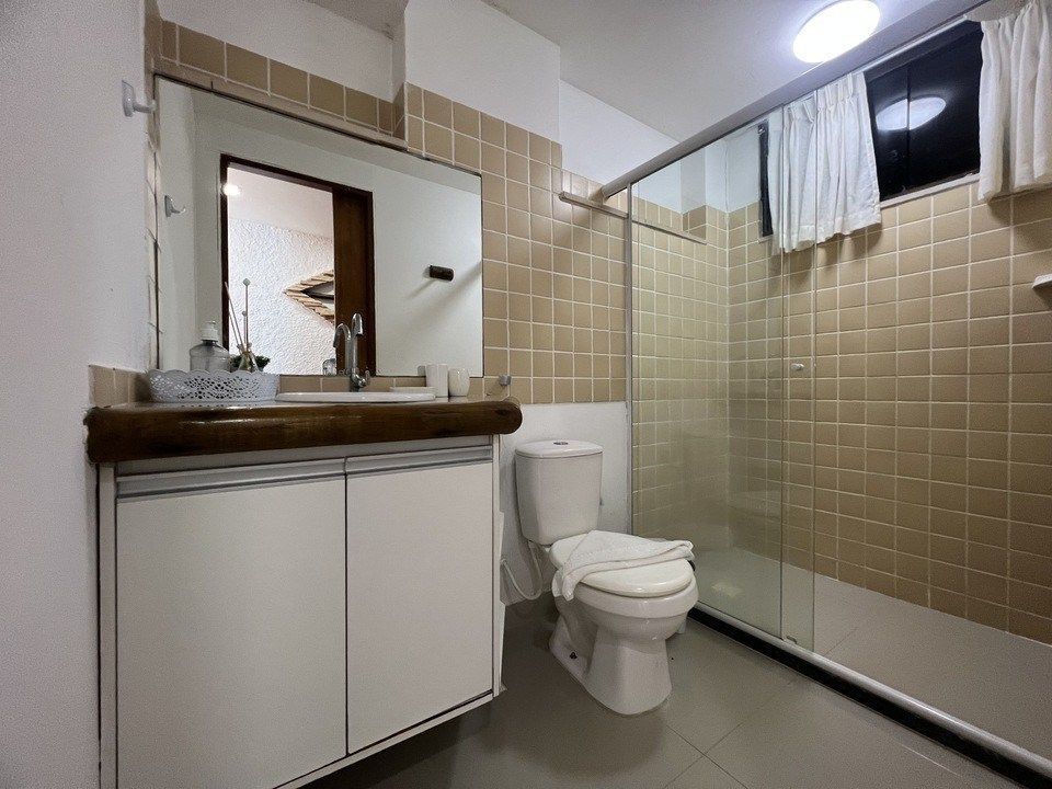 Banheiro com pia branca, vaso sanitário, box de vidro e paredes revestidas com azulejos bege.