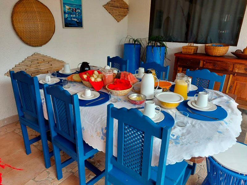 Mesa de café da manhã posta com cadeiras azuis, comida, bebidas e toalha de mesa branca em uma sala iluminada.
