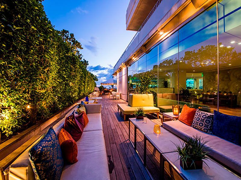 Lounge na cobertura ao entardecer com sofás, almofadas e uma parede de vidro; plantas verdejantes e iluminação ambiente.