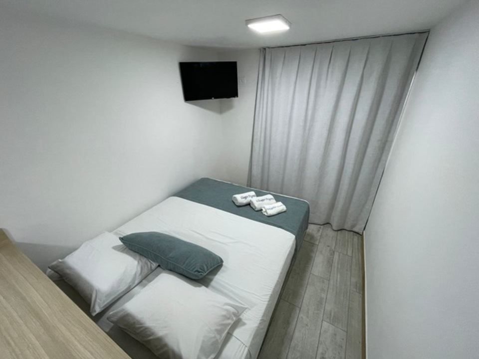 Quarto pequeno e minimalista com cama, TV e janela com cortina. Esquema de cores em tons de cinza claro e branco.