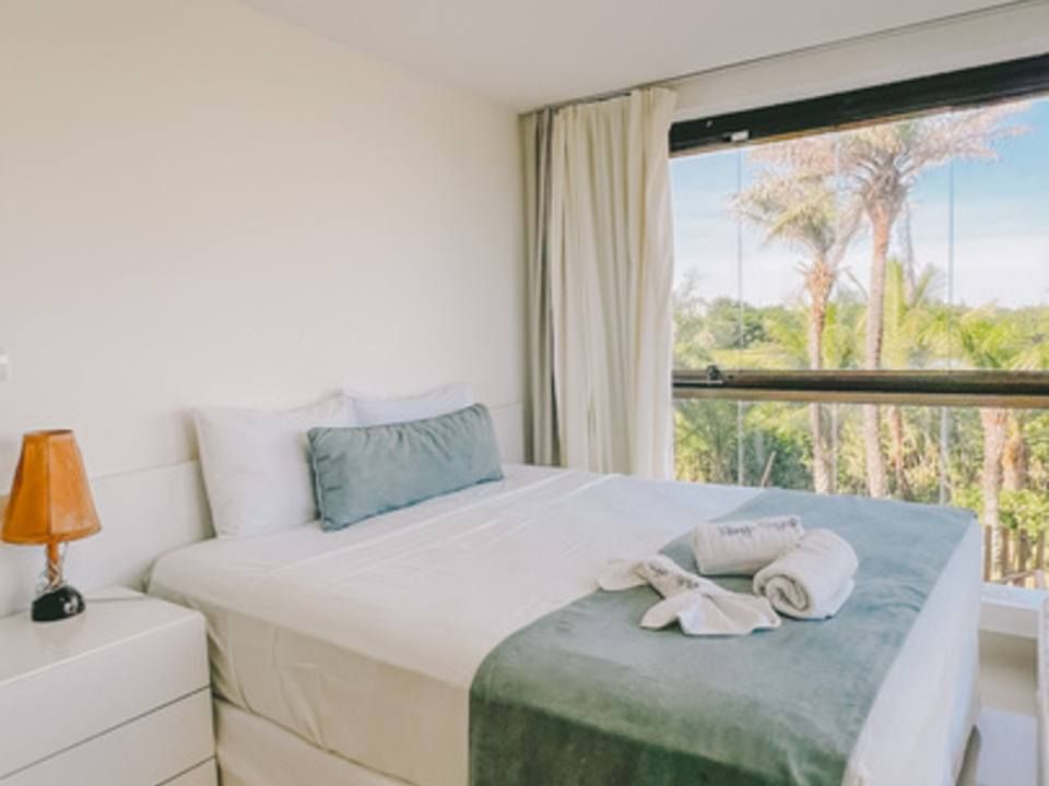 Quarto com paredes brancas, cama com detalhes em verde-azulado e uma grande janela com vista para as árvores.