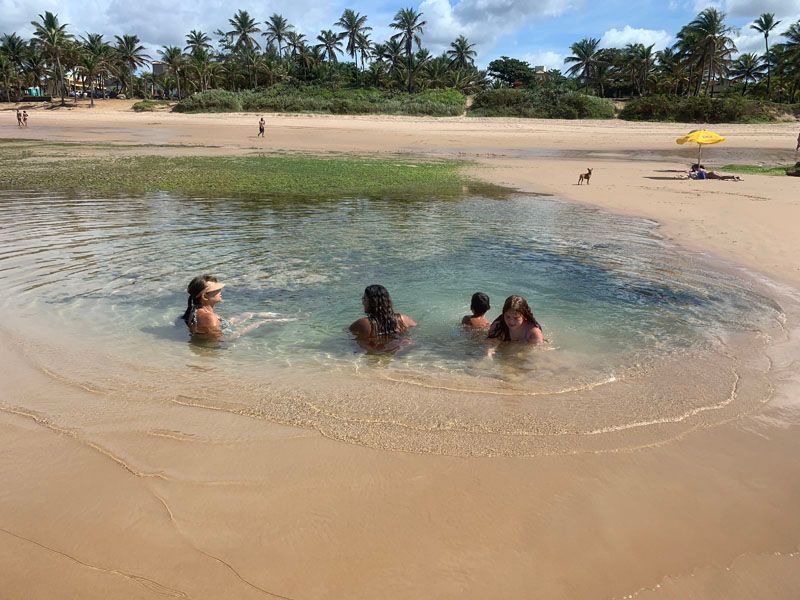 Pessoas nadando em uma poça de maré em uma praia de areia. Palmeiras, grama e um guarda-sol amarelo são visíveis.