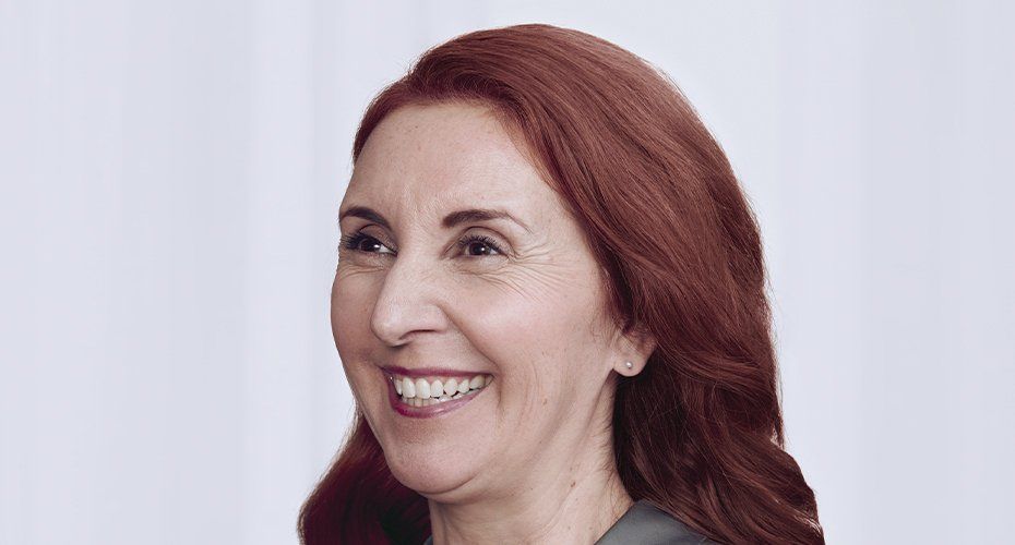 Woman with auburn hair smiles broadly, wearing a gray blazer against a light background.