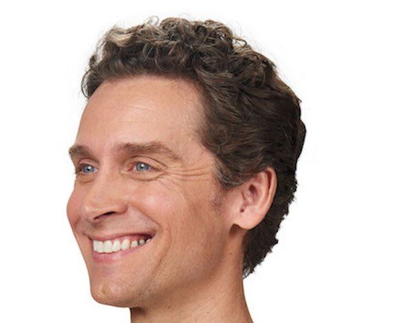 Man with curly brown hair smiles, showing teeth, against a white background.