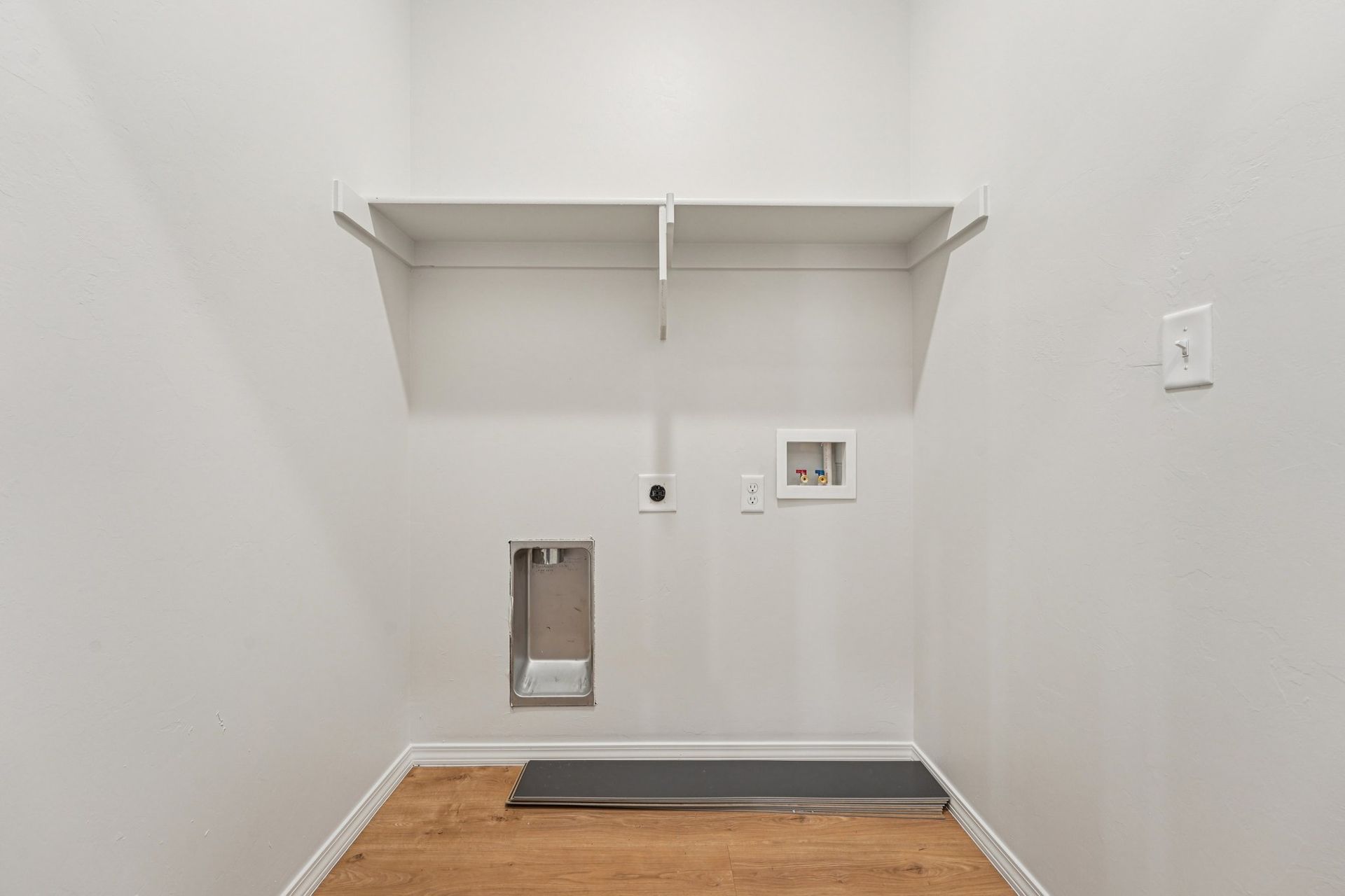 Empty, white-walled laundry room with wooden floor, shelf, and washer/dryer hookups.