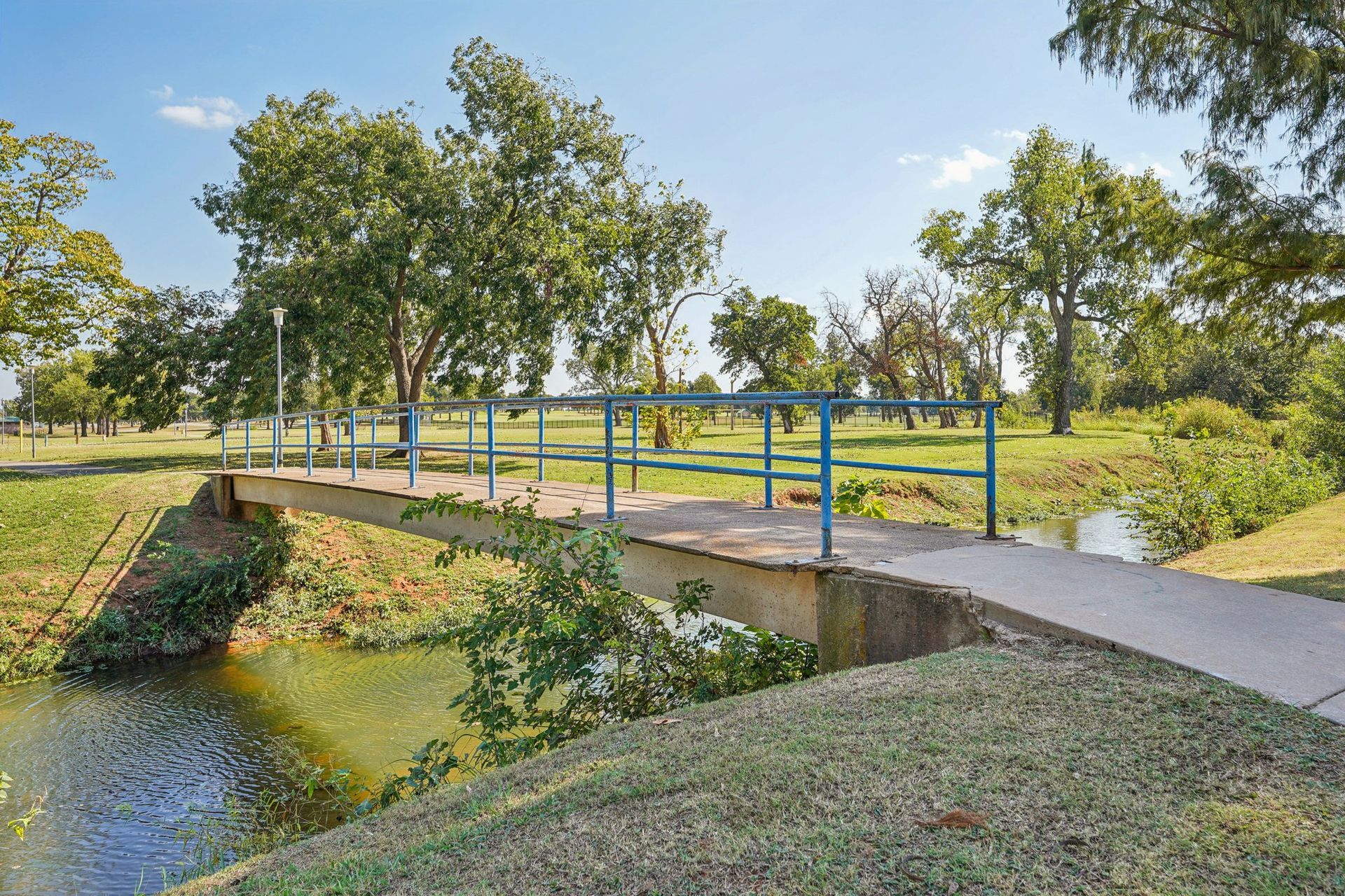 Concrete bridge over a waterway in a park, blue and green railings, sunny day.