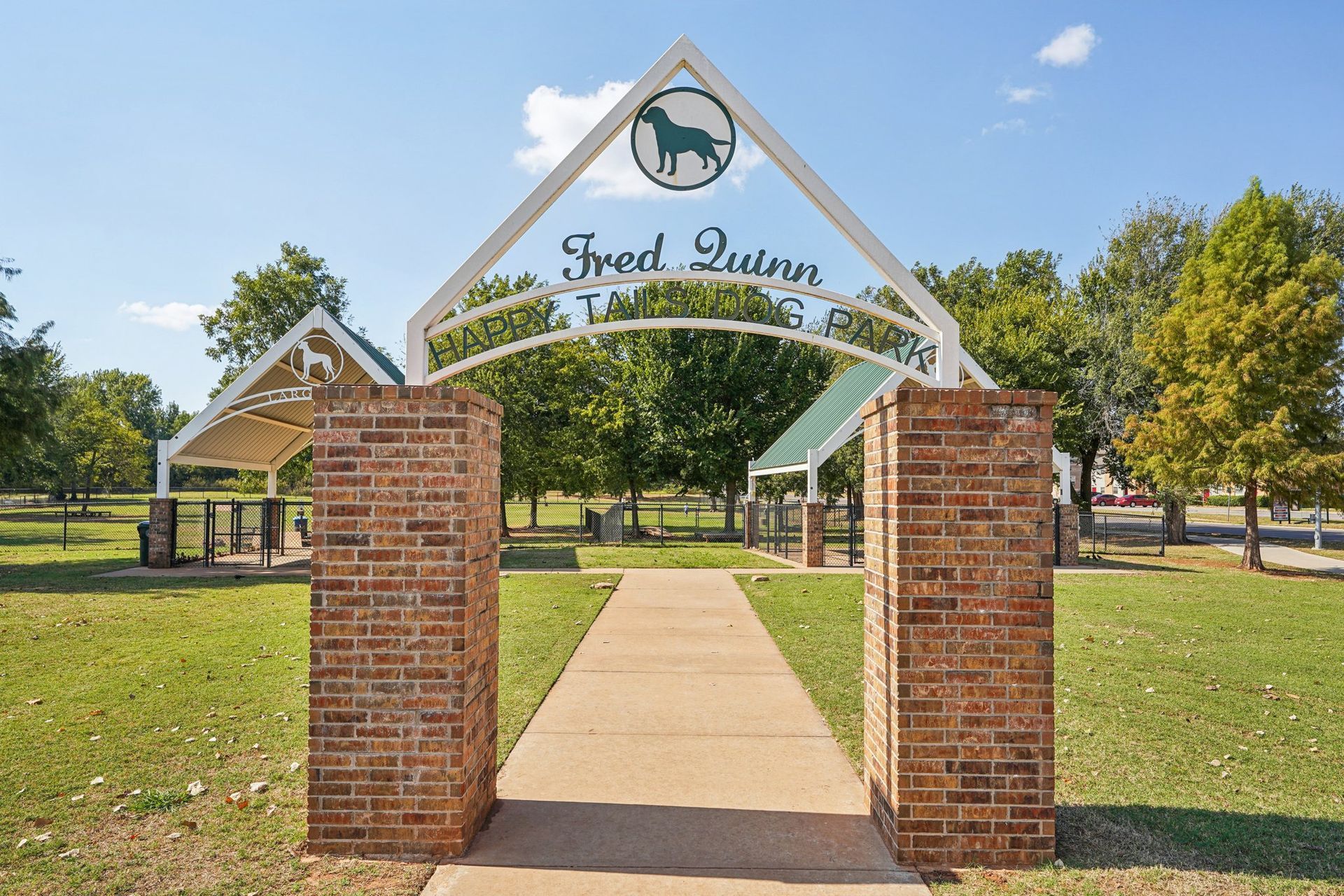 Arched entrance to Fred Quinn dog park; brick columns, paved path, green space, blue sky, and trees.