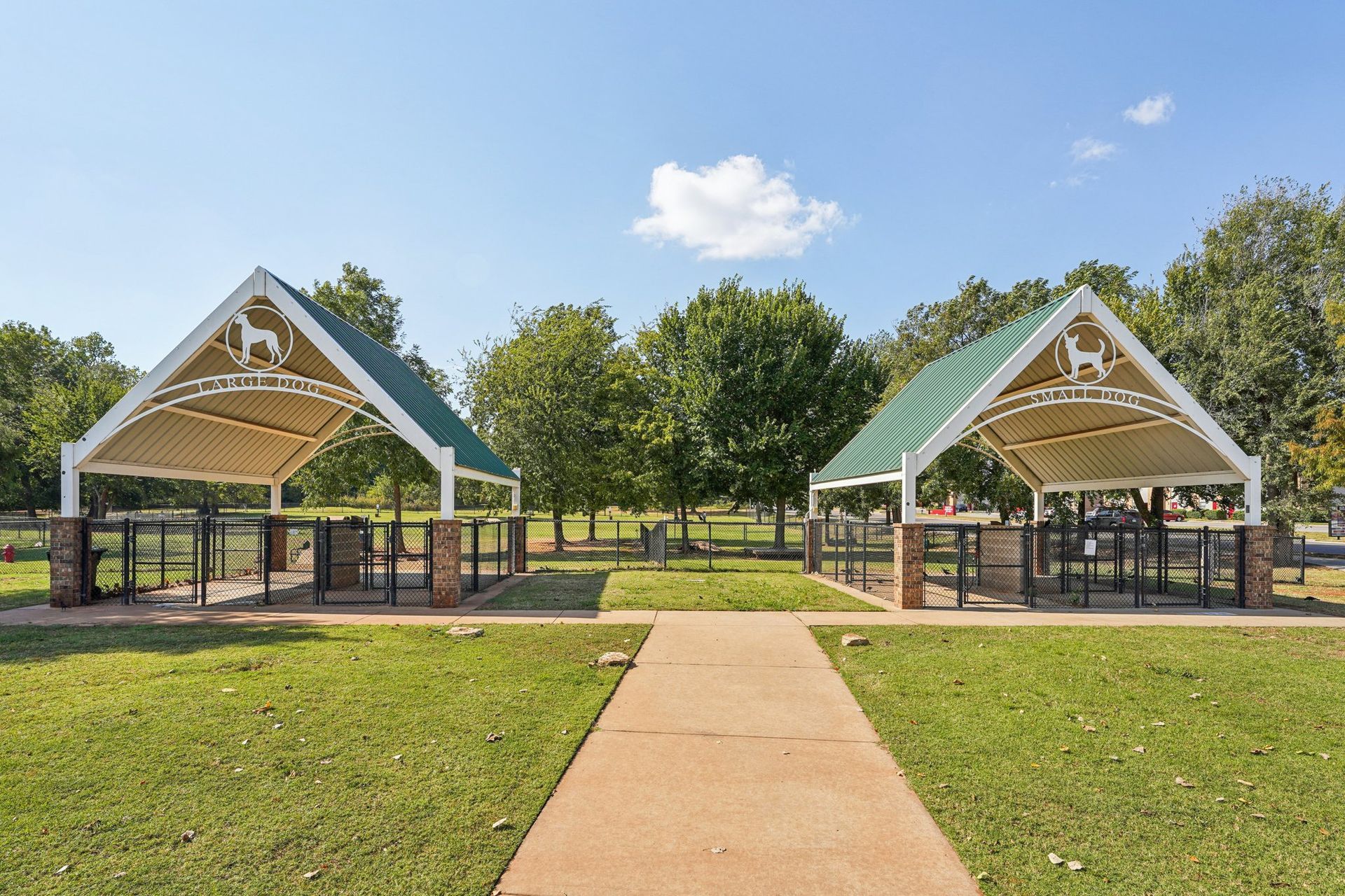 Two covered dog park entrances with green roofs, white frames, brown brick bases, and a pathway.