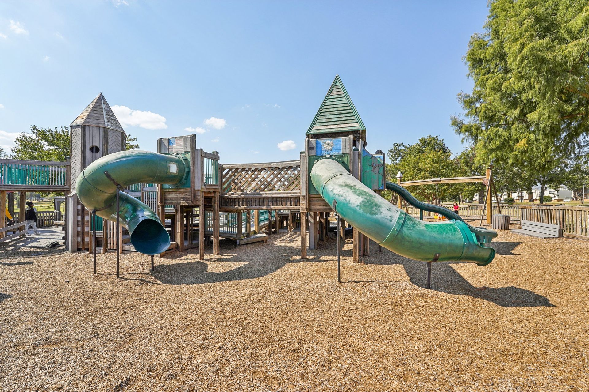 Playground with wooden structures, green slides, and wood chip ground cover under a blue sky.