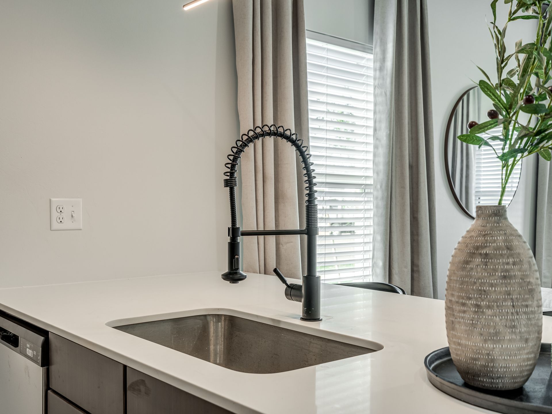 Kitchen with black faucet, stainless steel sink, white countertop, and gray curtains.
