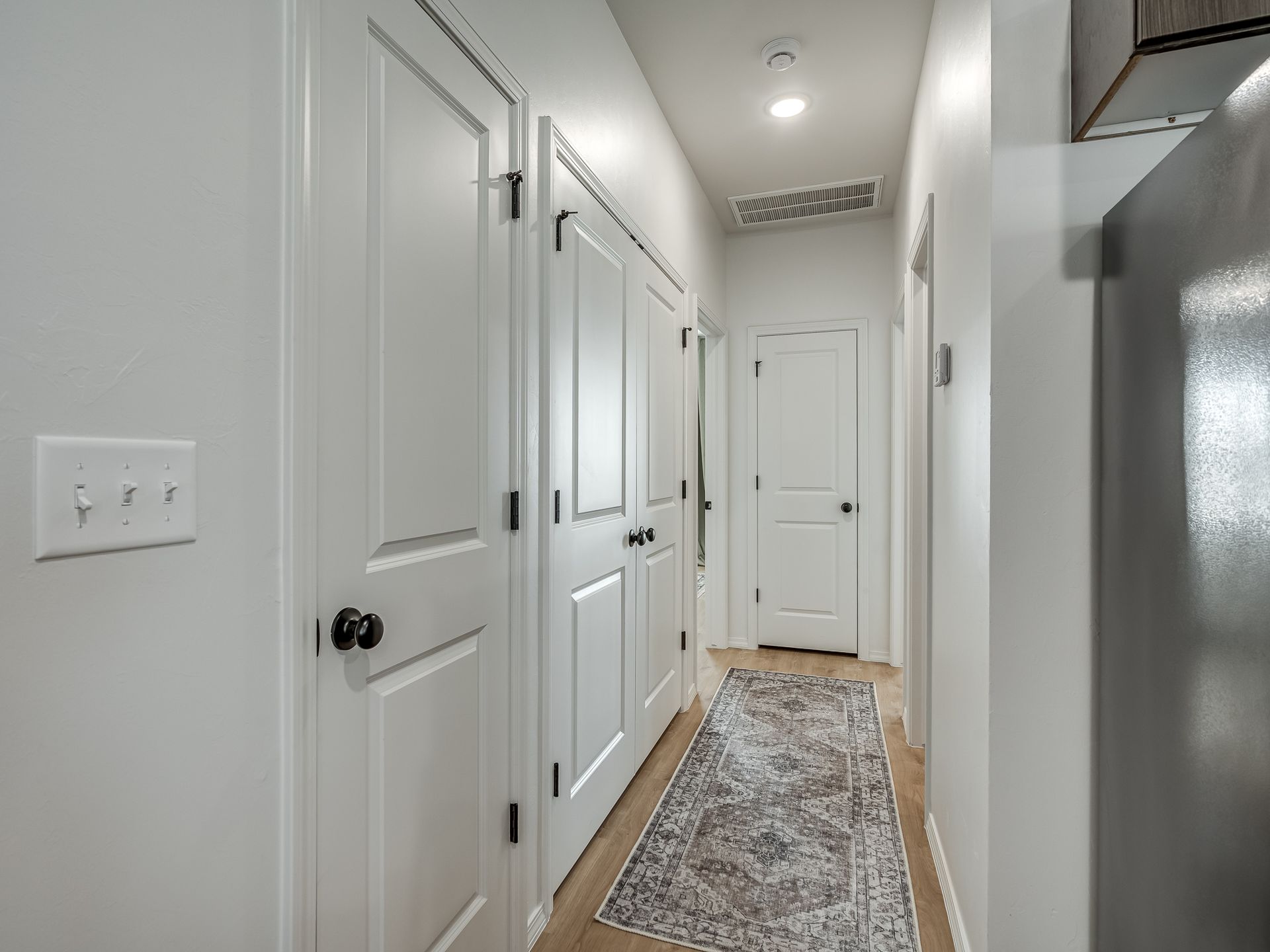 Narrow hallway with white doors, light wood floor, and patterned rug.