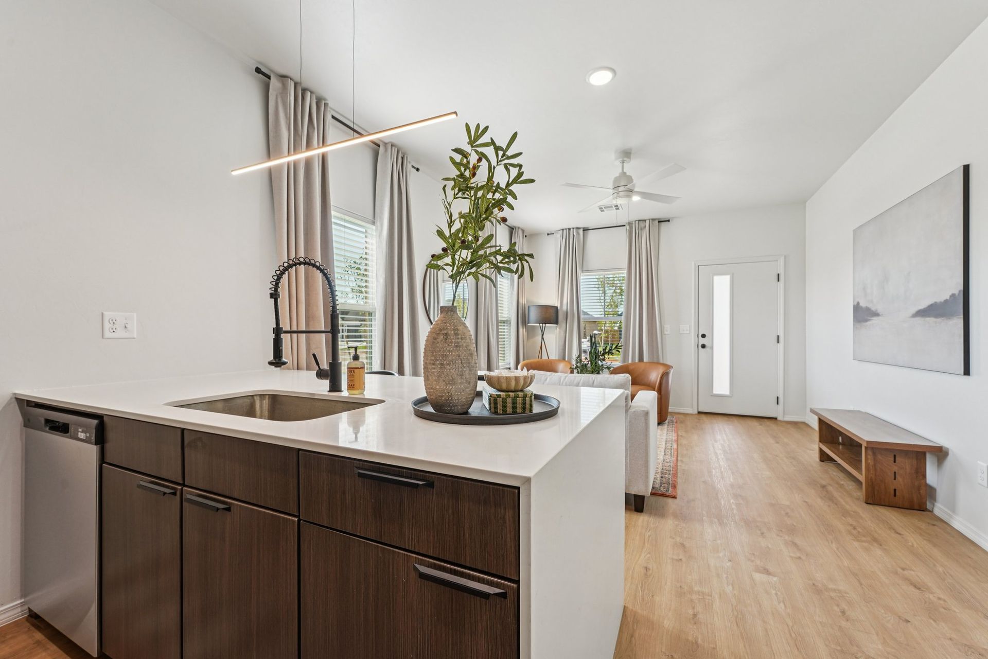 Modern kitchen with island, sink, and wood cabinets, opening to a living area with a bench and artwork.