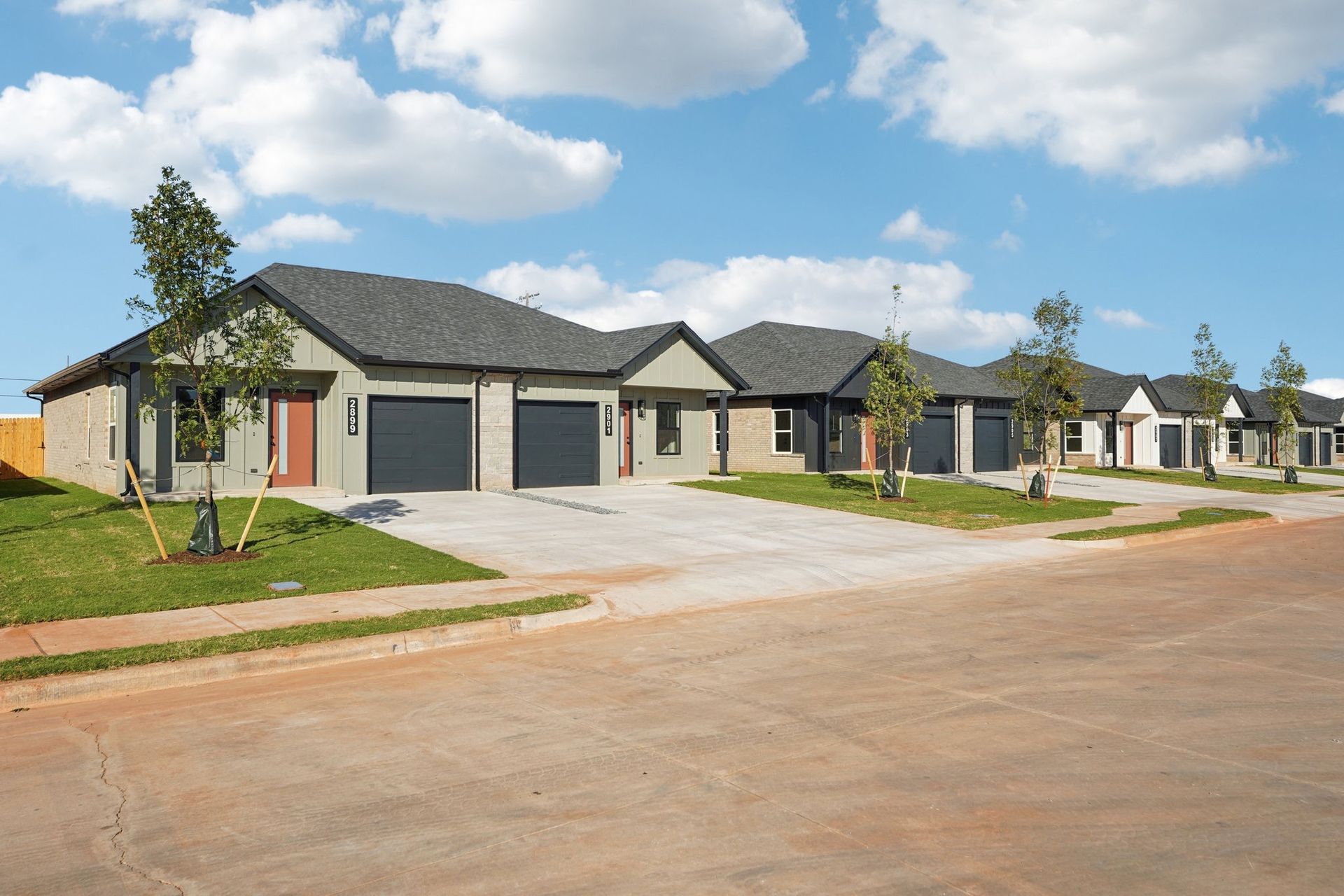 Row of modern, single-story homes with attached garages, gray roofs, and brown and green landscaping under a blue sky.