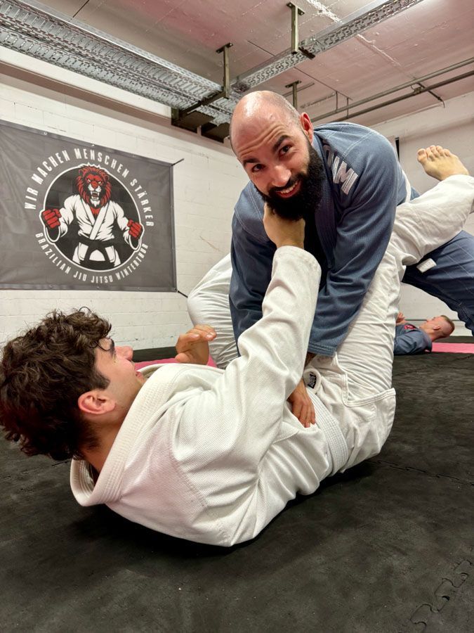 Ein Mann mit Bart bringt einem jungen Mann in einem Fitnessstudio Jiu-Jitsu bei.